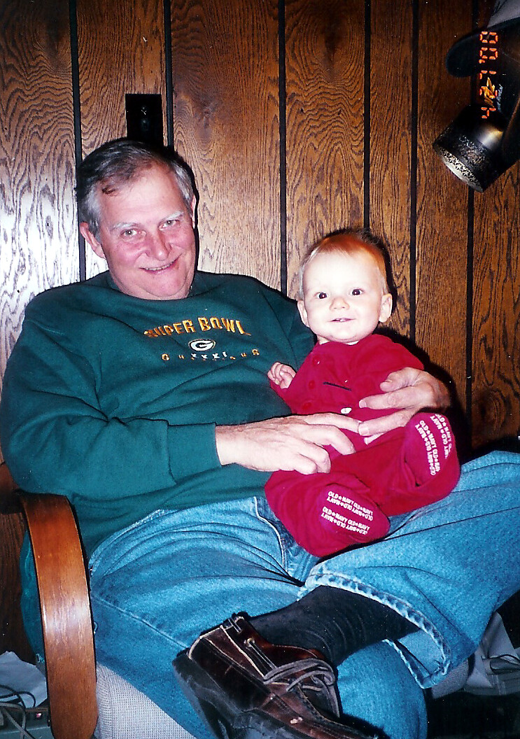 Older man in a green sweatshirt sits in a chair, holding a smiling baby in a red outfit on his lap in a wood-paneled room.