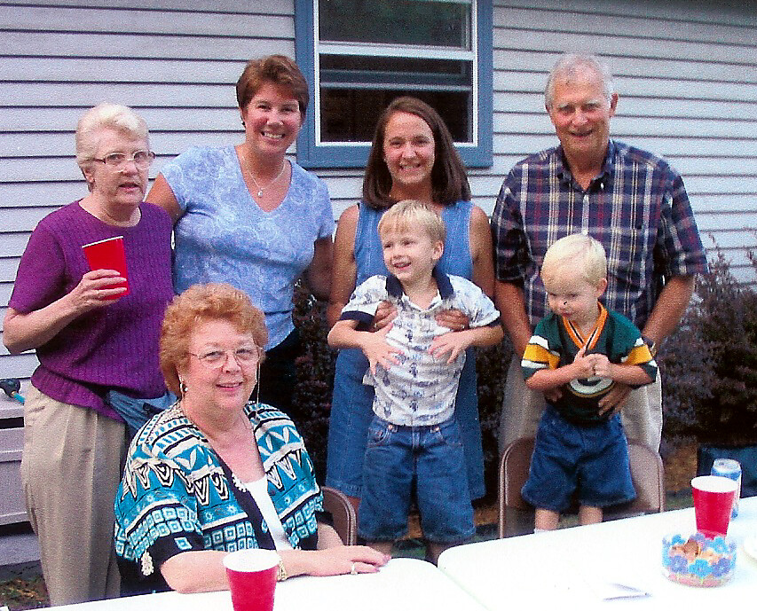 Family group photo in a backyard: three generations standing behind a seated woman at a table with red cups and a small cake.