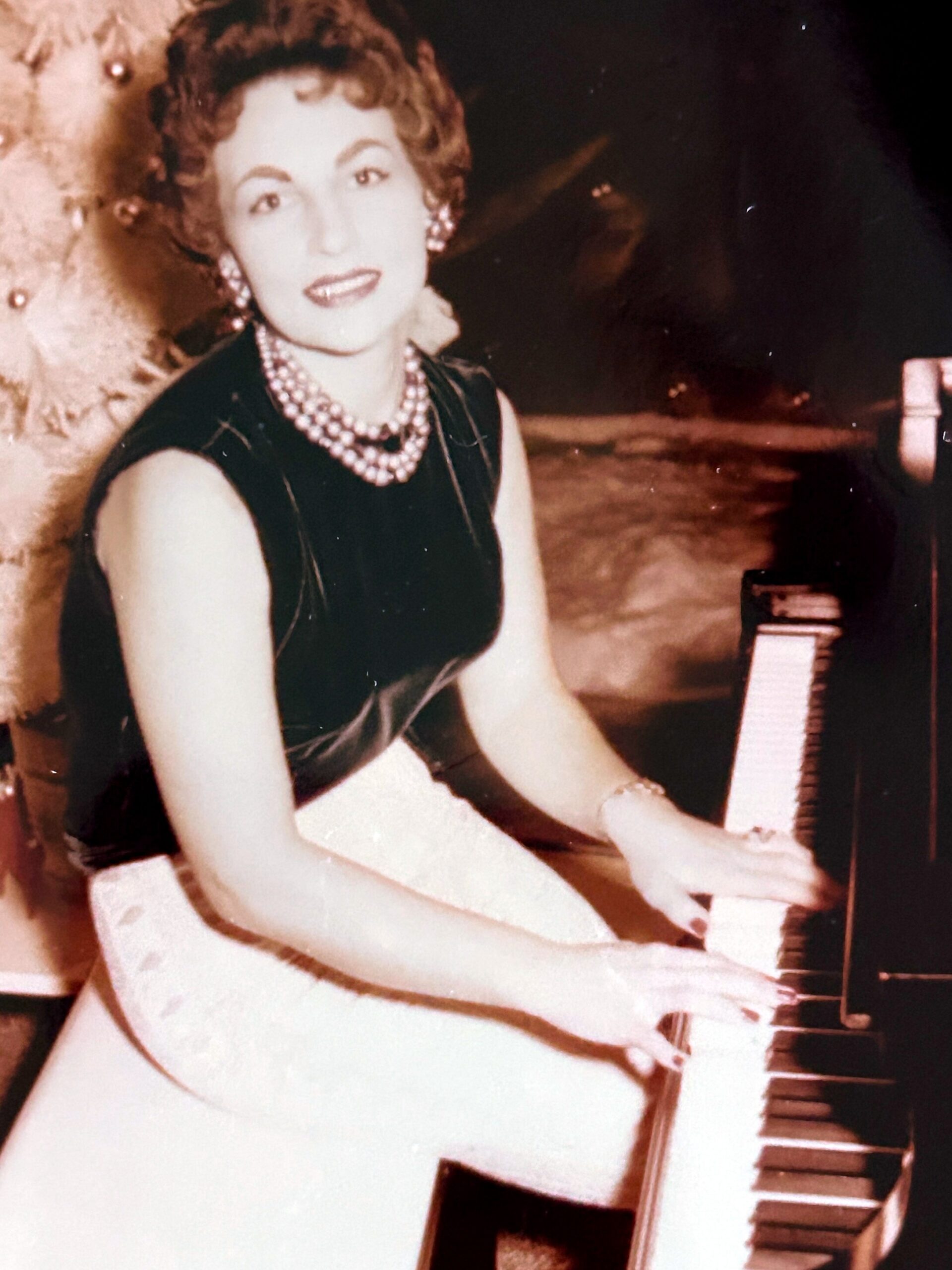 Woman in a black sleeveless dress with a pearl necklace sits beside a white guitar, looking at the camera.