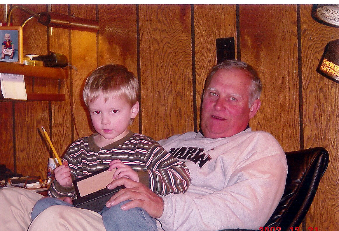 Young boy sits on an older man’s lap, holding a pencil and a small box in a wood-paneled room.