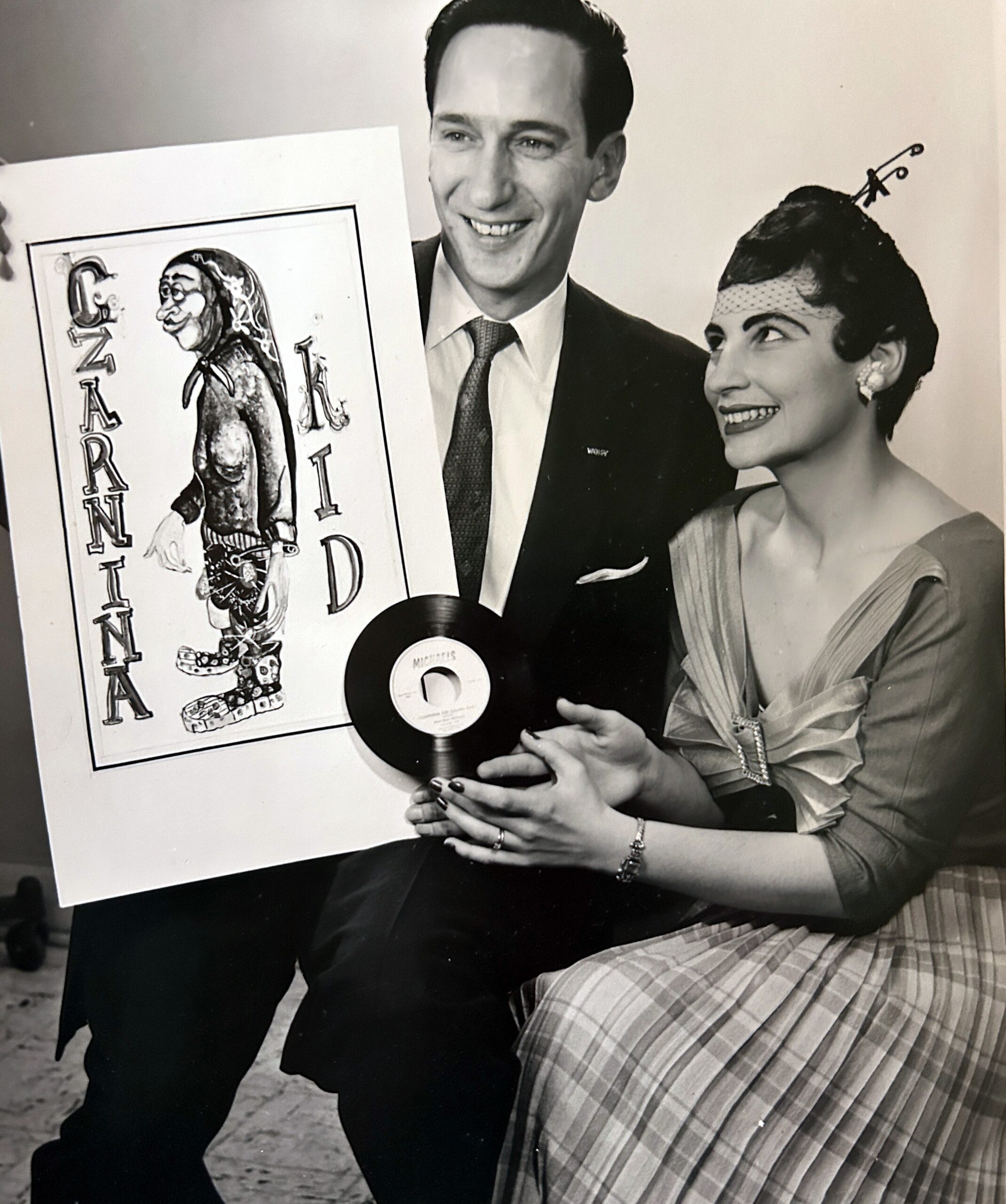 Black-and-white photo of a smiling man in a suit and a woman in a vintage dress posing with a large illustrated poster and a vinyl record posed between them.