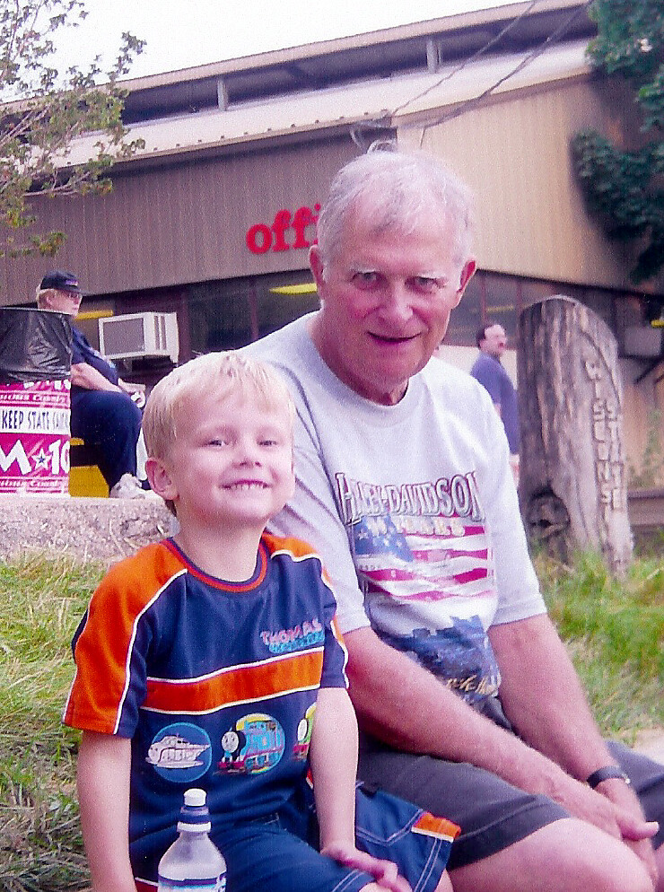 Grandfather and grandson sit on a curb, smiling at the camera outside a building with an 'office' sign.