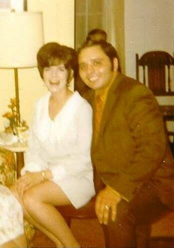 Smiling woman in a white dress sits on a chair beside a man in a brown jacket in a living room, posing for a photo with a lamp and chairs in the background.