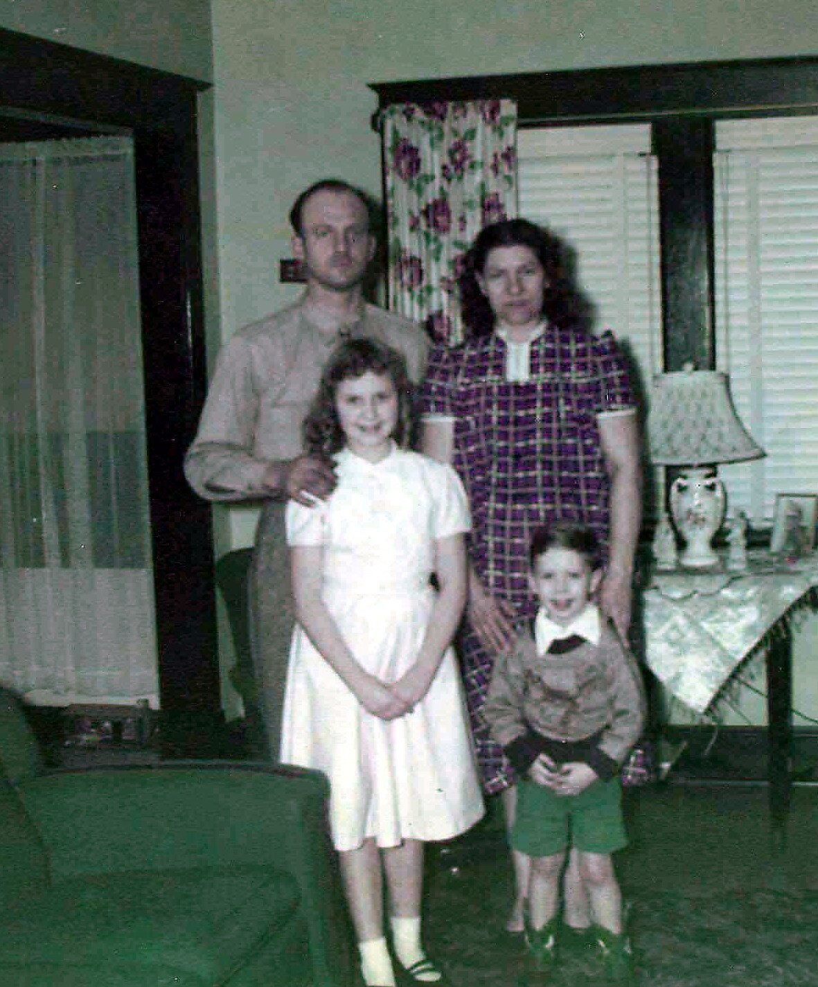 Family of four posing for a photo in a living room: father with hand on daughter’s shoulder, mother beside, plus a young boy in front.