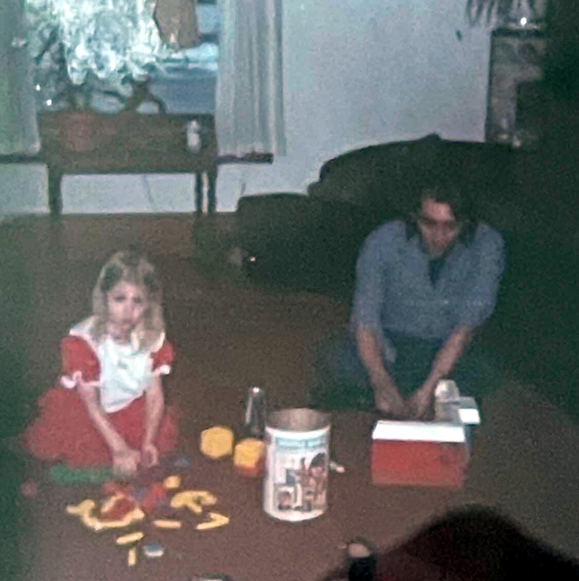 Two people kneeling on the floor around a low table with snacks and a bucket, girl in red dress on the left and man in gray shirt on the right.