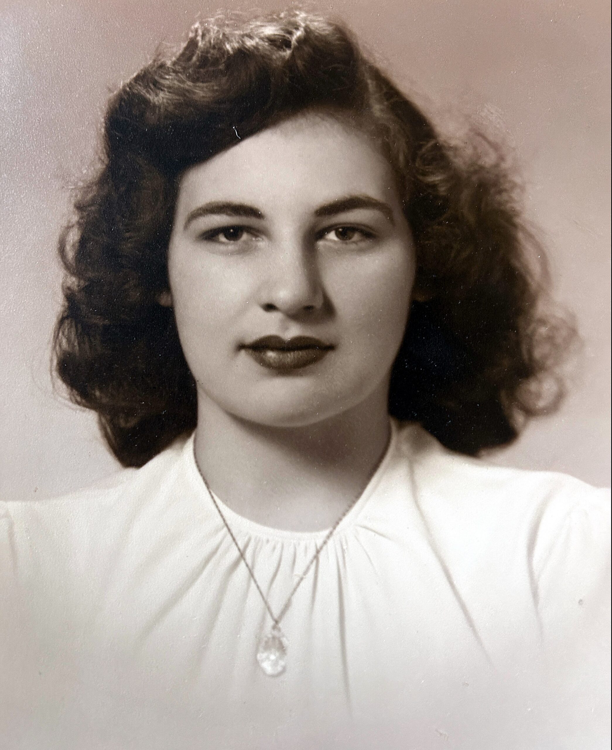 Vintage black-and-white portrait of a young woman with dark, wavy hair and a teardrop pendant necklace wearing a light blouse, facing the camera.