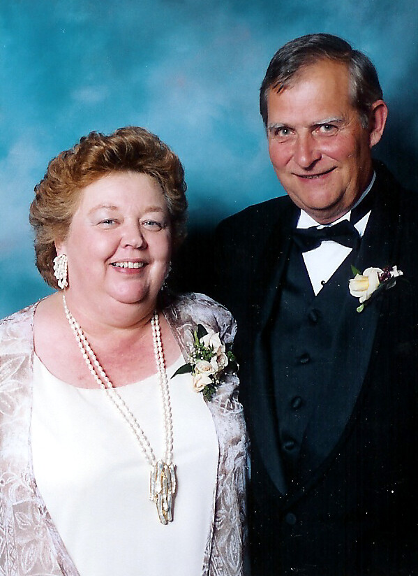 Smiling older couple posing in a formal studio portrait; woman in a white dress with a lace shawl and pearl necklace, man in a black tuxedo with boutonniere.