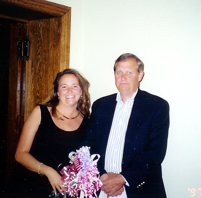 A smiling woman in a black dress standing beside a man in a dark blazer, holding a pink ribbon bouquet together in a doorway.