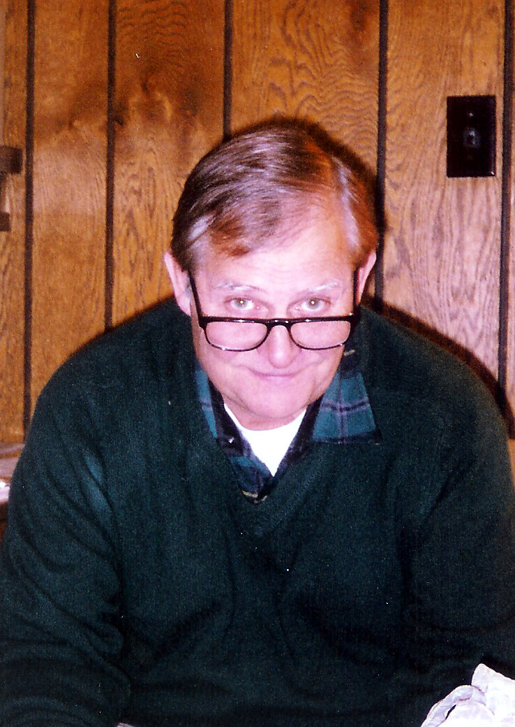 Man with glasses leaning forward, looking at the camera, wearing a dark green sweater with a plaid collar against wood-paneled walls behind him,