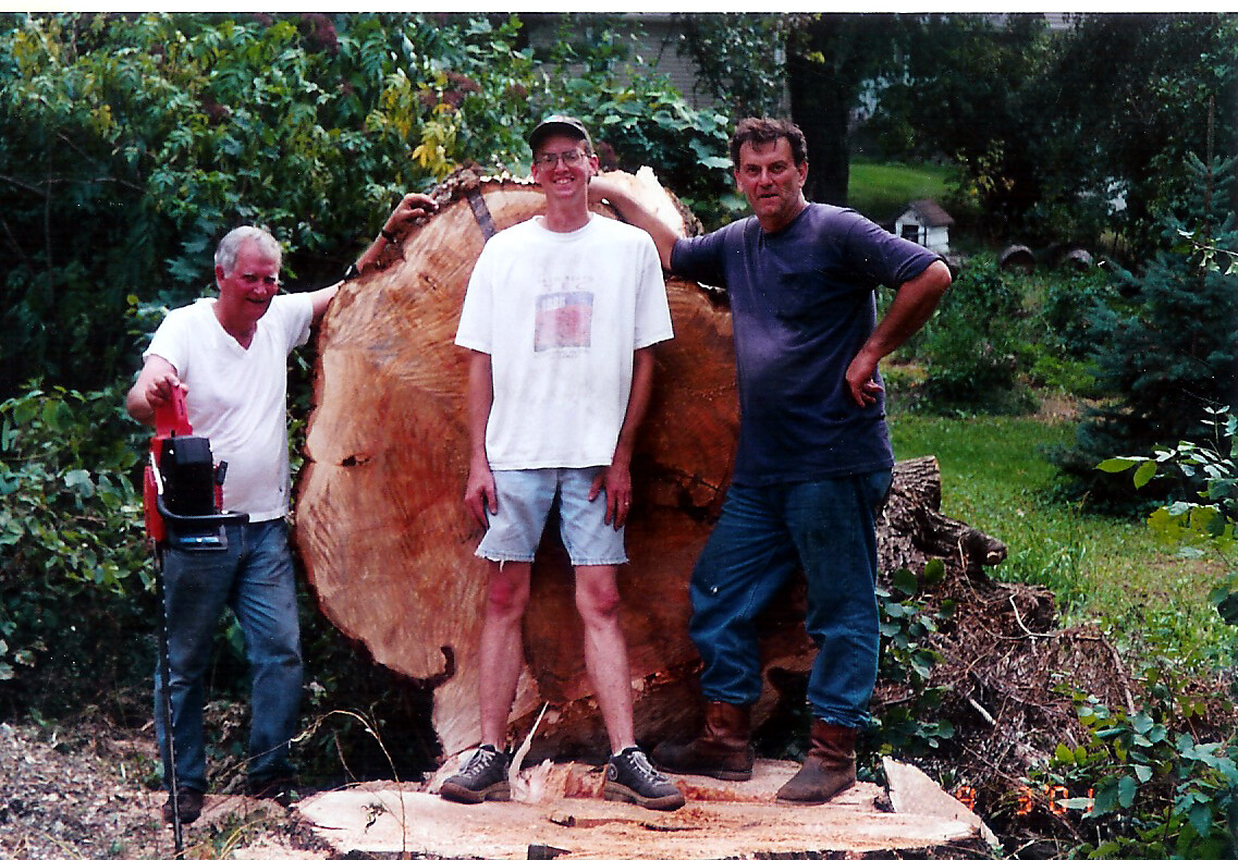 Three men pose in a backyard beside a huge tree stump after cutting it down.