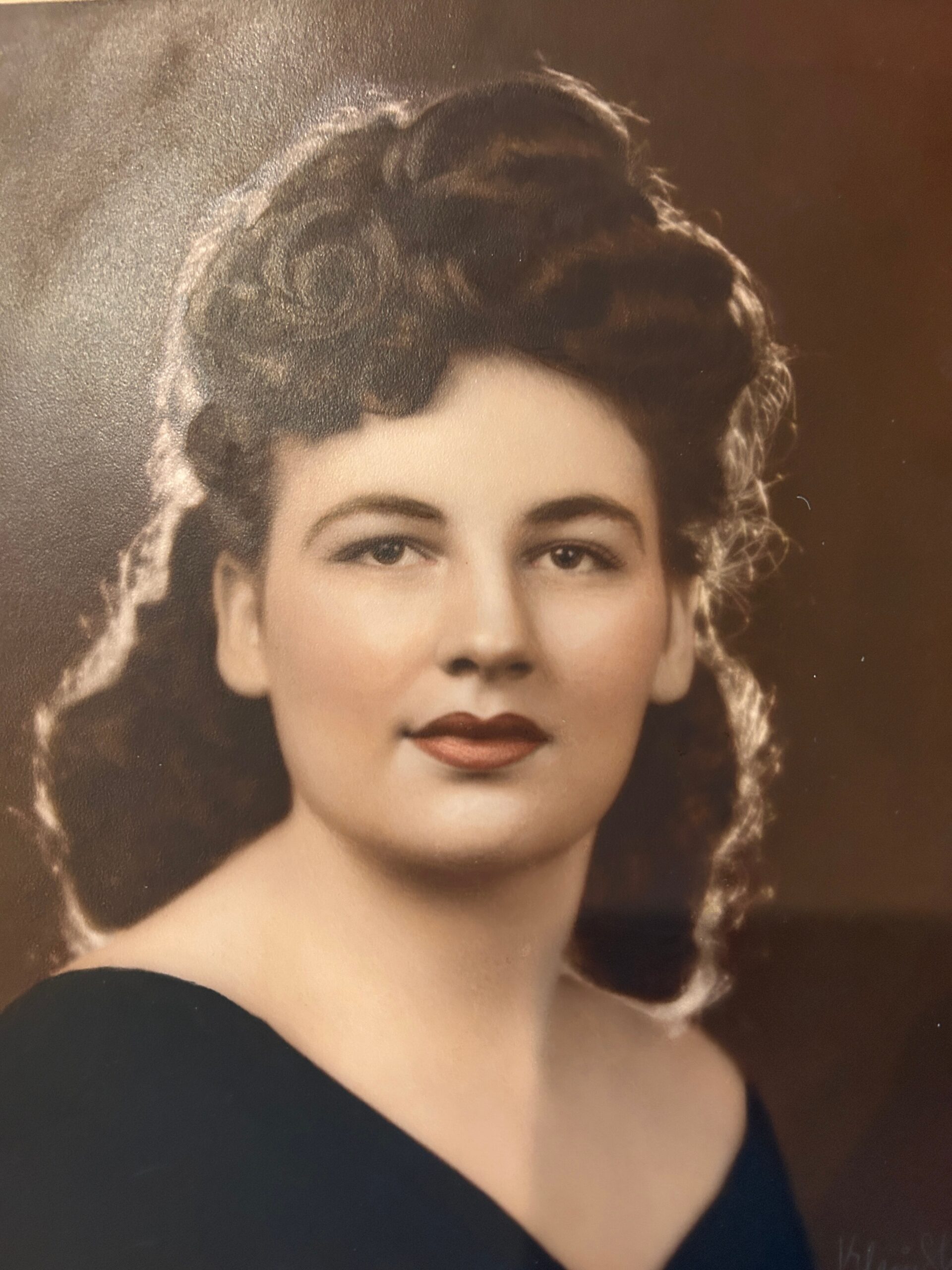 Vintage studio portrait of a young woman with dark, styled hair and subtle makeup, wearing a dark off-the-shoulder dress against a brown backdrop.