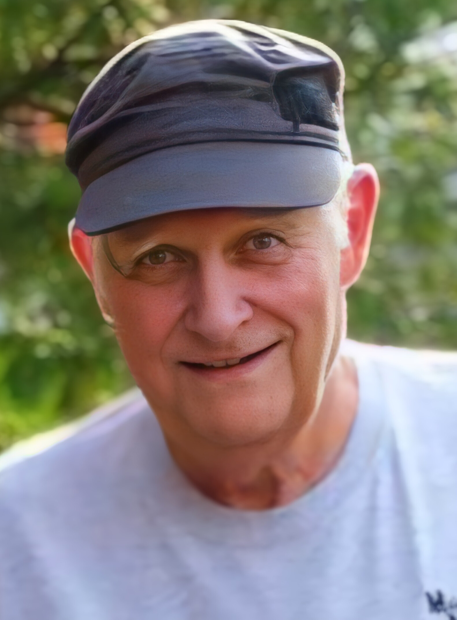 Close-up of a smiling man wearing a gray cap with sunglasses resting on the brim, outdoors with greenery in the background.