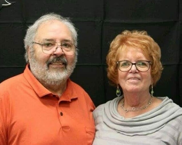 Two smiling older adults posing together, man in an orange polo shirt with a gray beard and glasses, woman with short red hair in a light gray top and glasses against a dark backdrop.