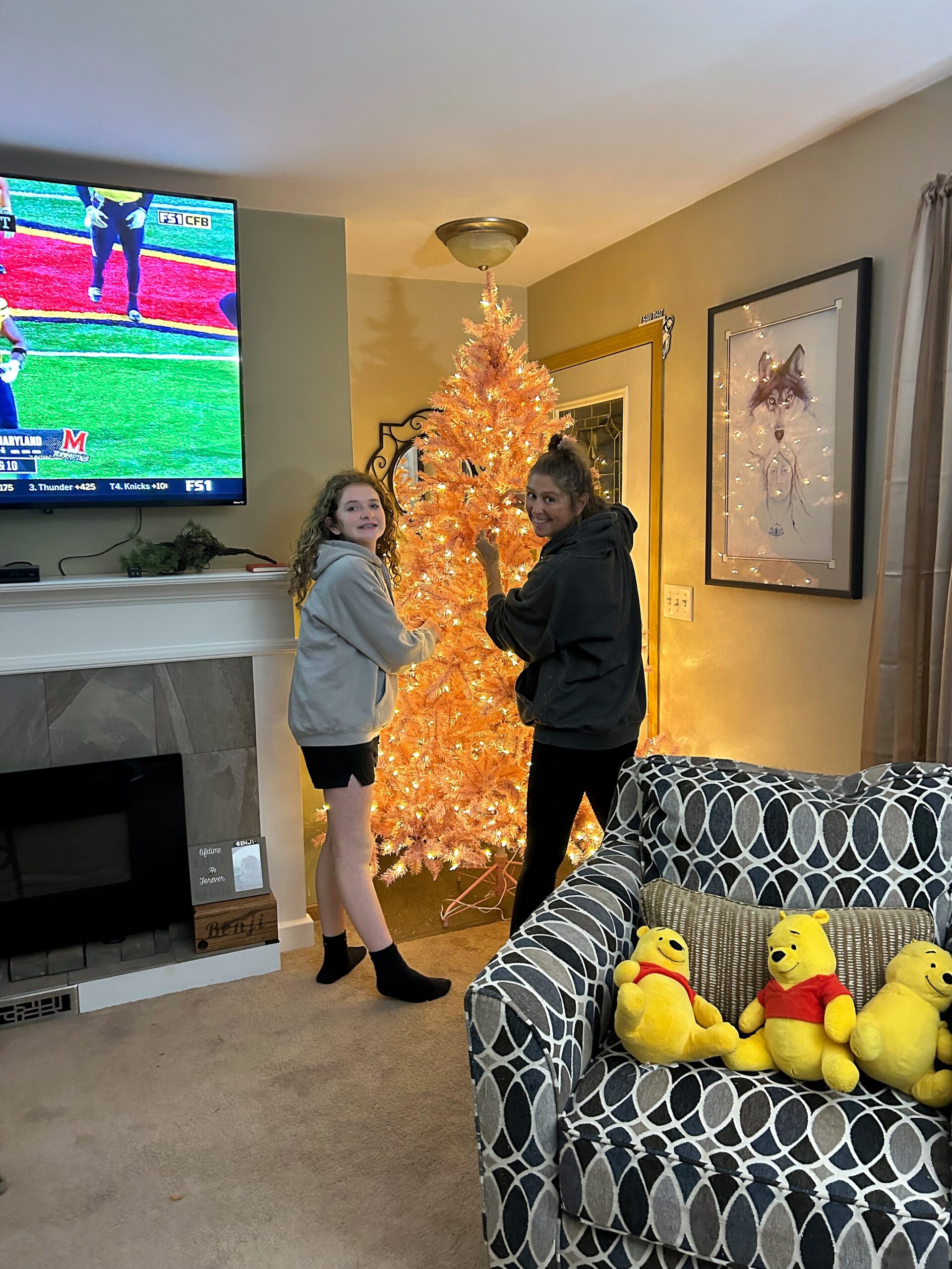 Two people stand by a pink-lit Christmas tree in a cozy living room, smiling at the camera.
