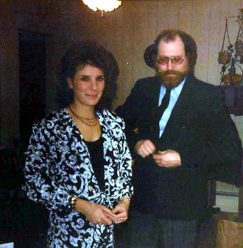 Woman in a black-and-white patterned blazer and necklace posing with a bearded man in a suit indoors at a social event.