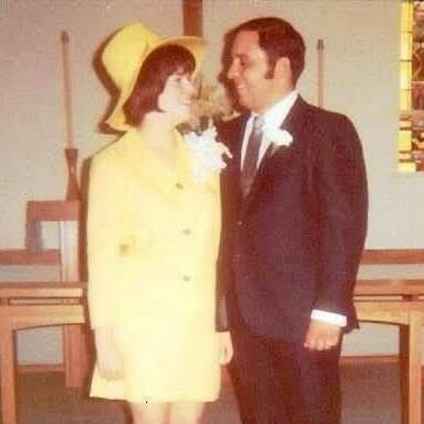 Smiling couple in a yellow dress and hat and a man in a dark suit with a boutonniere, standing close together in a church-like room.