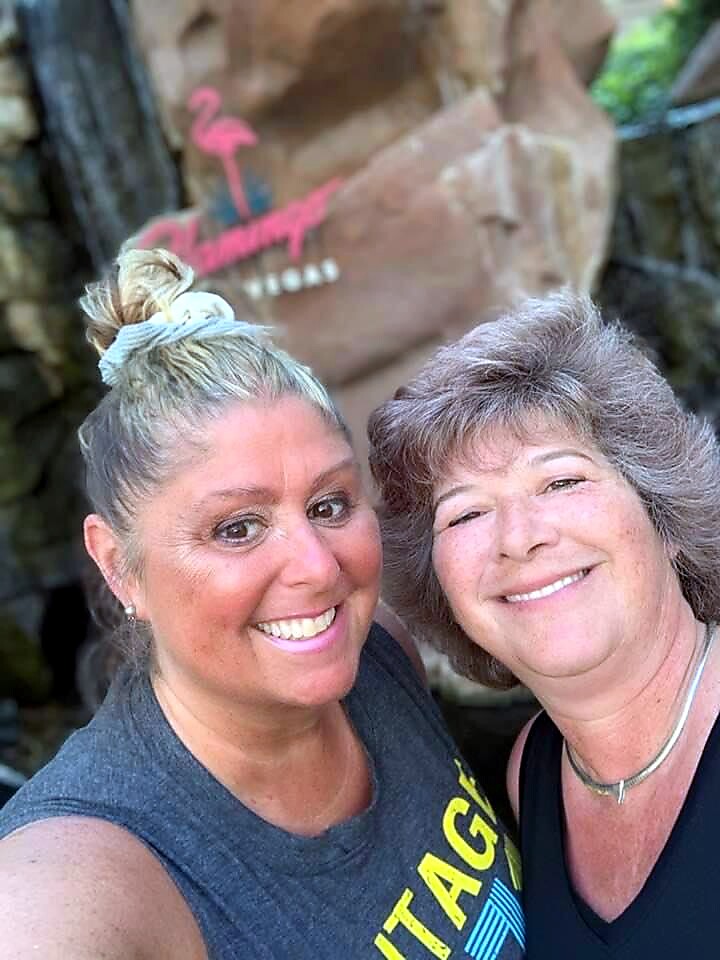 Two smiling women take a selfie outdoors, with a rocky waterfall behind and a pink flamingo sign in the background.