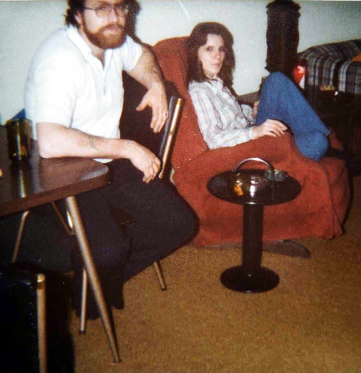 Man with a beard sits at a table beside a red sofa while a woman in a striped shirt lounges with legs on the sofa in a living room scene