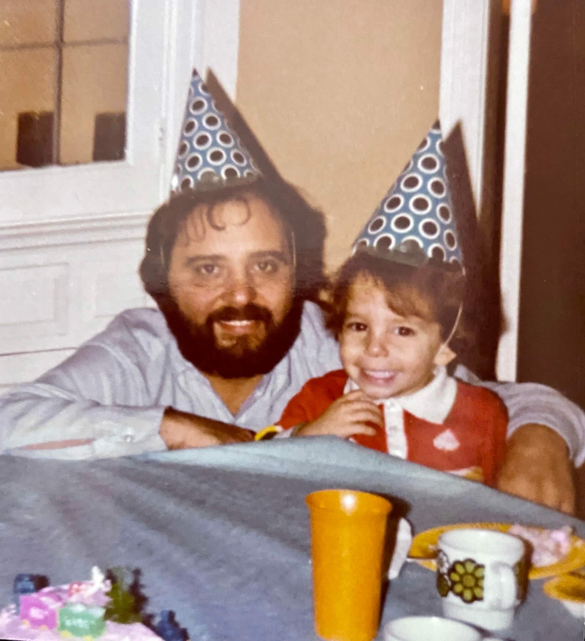 Man with beard and child wearing blue polka-dot party hats, smiling at a birthday celebration group photo