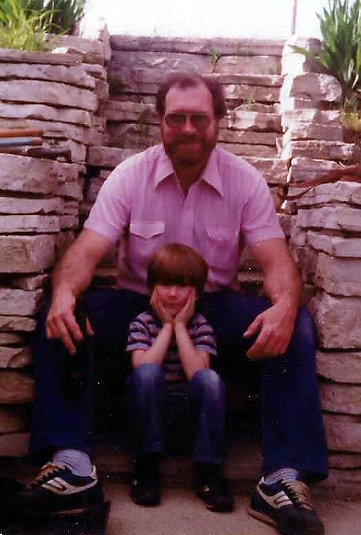 Man in a pink shirt sits with a small child between his legs on a stone stair wall outdoors.
