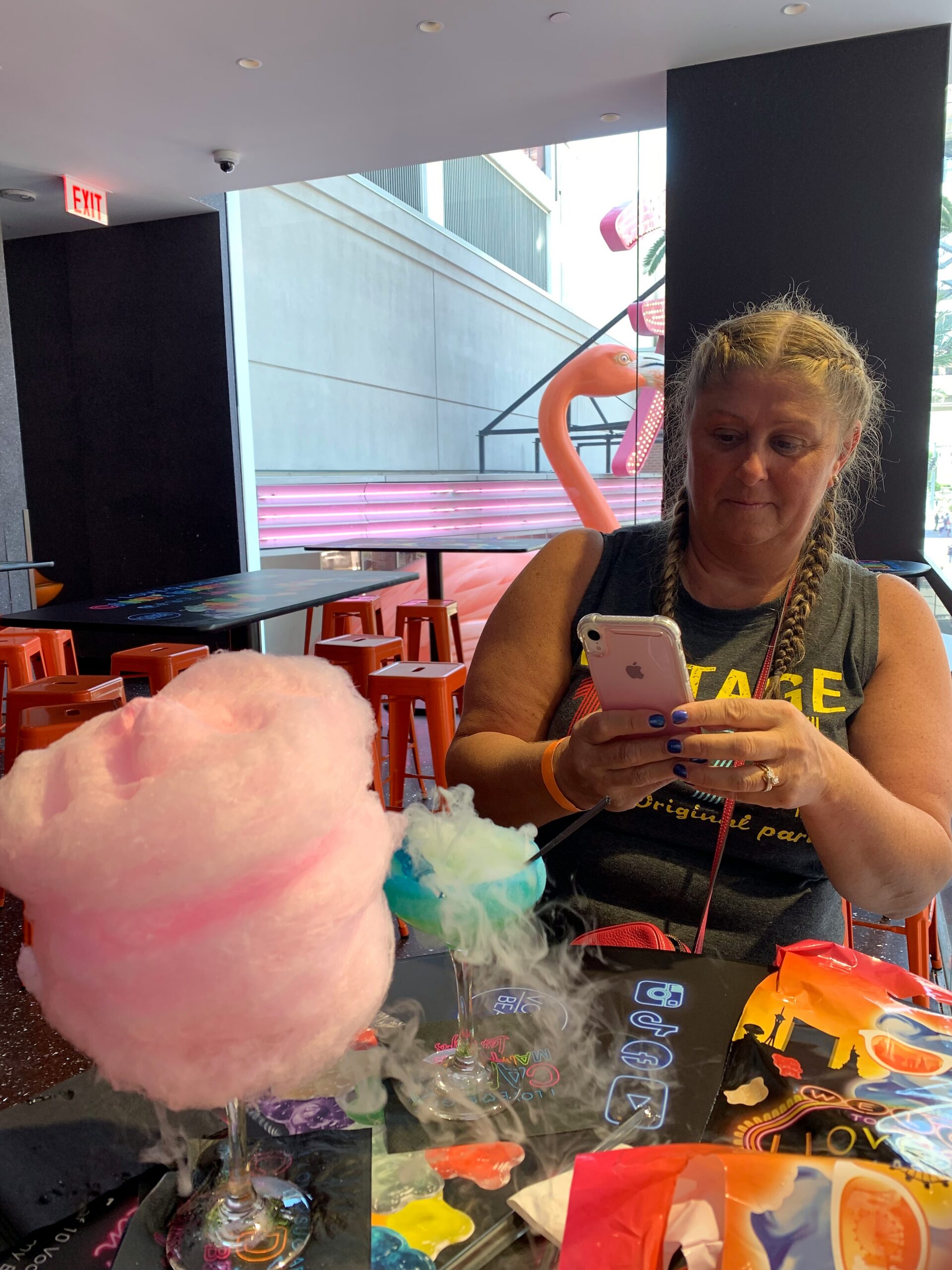 Woman with braided hair looking at her phone while seated at a table with colorful candy and neon pink decorations visible outside the window.