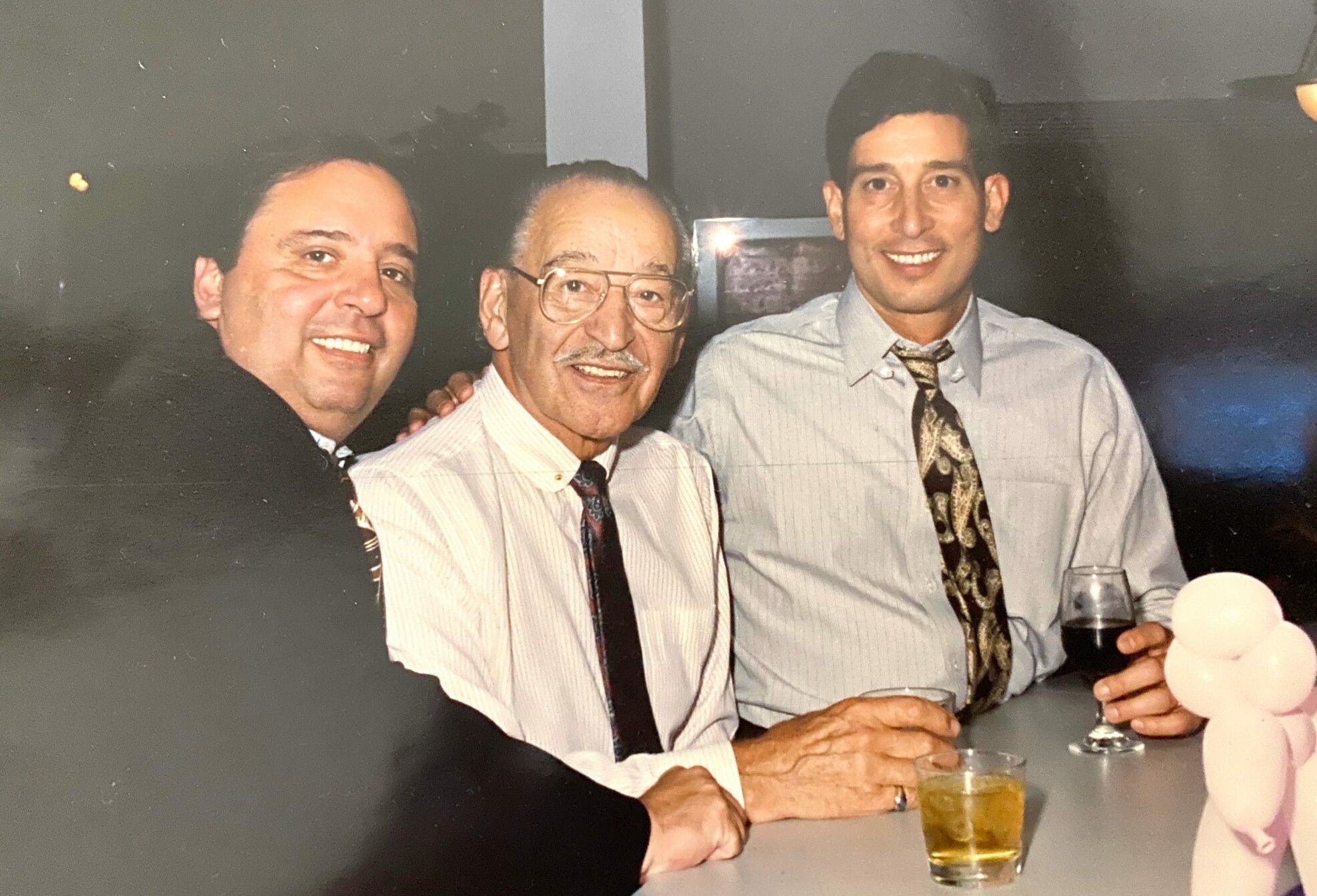 Three men in dress shirts and ties pose at a bar, smiling at the camera with drinks in hand.