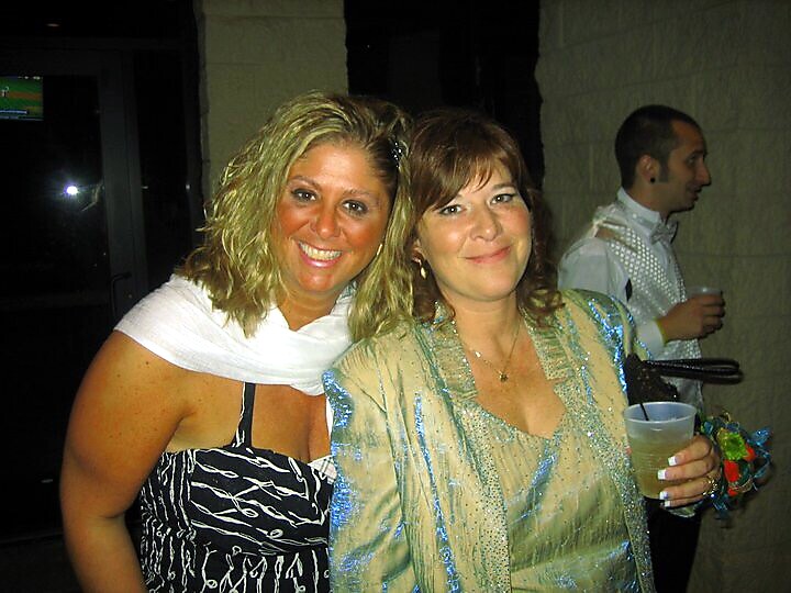 Two women posing for a photo at a party, smiling; one in a black patterned dress with a white scarf, the other in a light blue shiny top.