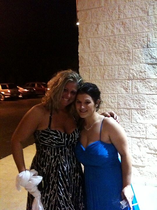 Two women in formal dresses pose together outside a light-colored brick wall, smiling with arms around each other at night.