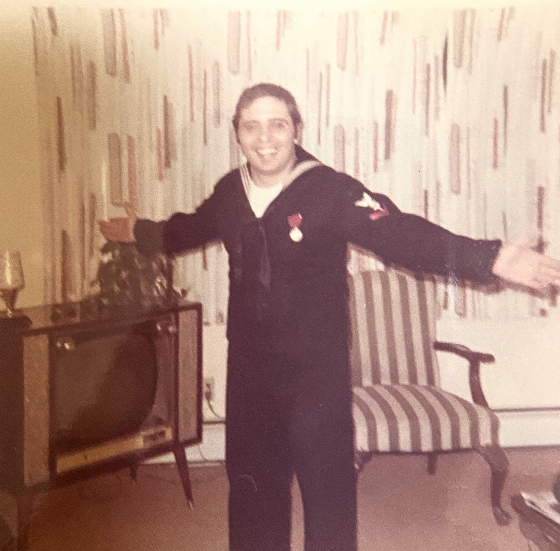 Person standing with arms outstretched in a living room, smiling at the camera, wearing a dark outfit. A striped armchair and cabinet are visible in the background.