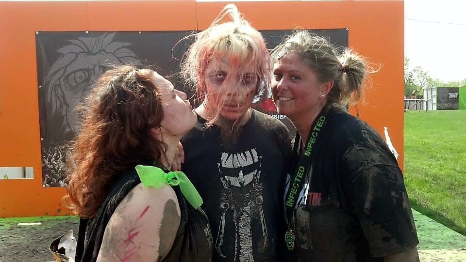 Three people posing for a photo at a zombie makeup event; central person has pale face and torn hair, flanked by two friends.