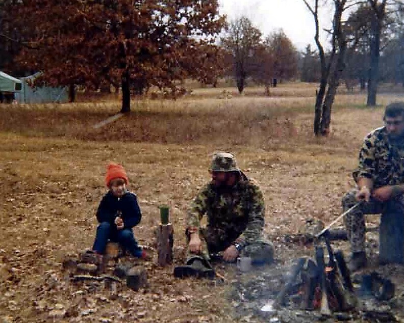 Child wearing an orange knit hat sits on logs near a small campfire while two adults in camouflage are nearby in a leaf-filled autumn field.