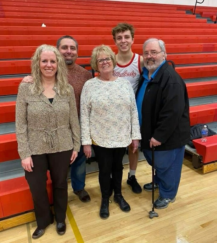 Five people pose for a group photo in a gym with orange bleachers behind them: a smiling blonde woman in a patterned blouse, a man in a brown sweater, an older woman in a white knit sweater with glasses, a tall young man in a basketball jersey, and an older man with a beard in a blue shirt and black jacket using a cane.