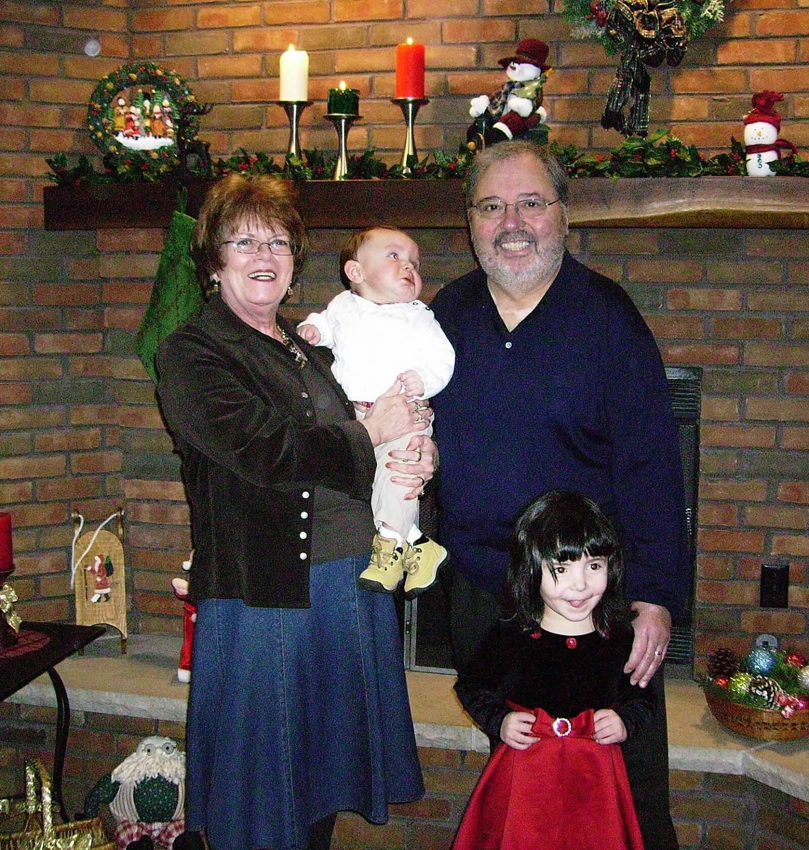 Grandmother and grandfather smile with two children—a baby and a young girl—in front of a brick fireplace decorated for Christmas with candles and greenery.