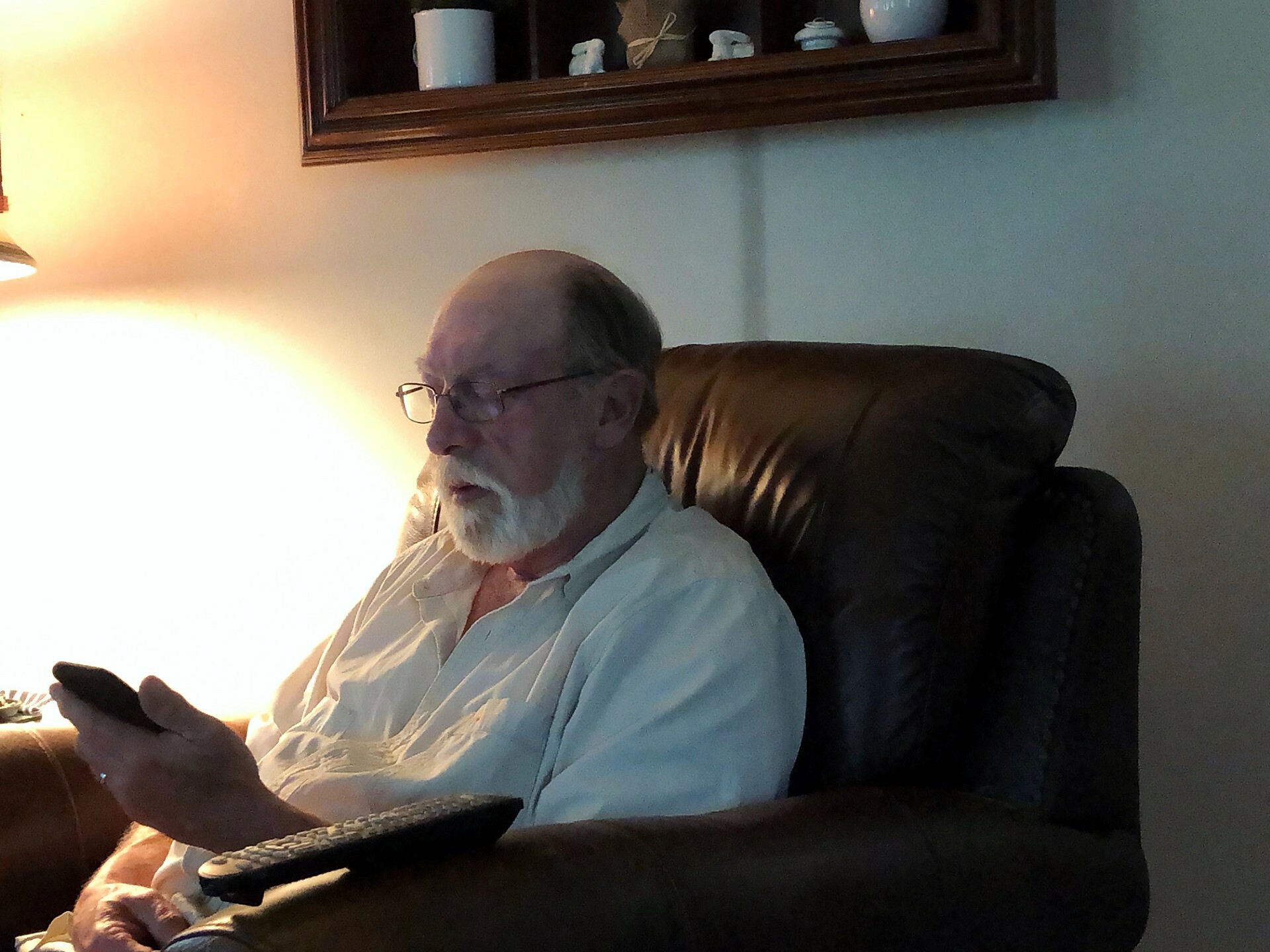 Older man with glasses and white beard sitting in a brown recliner, looking at a smartphone in his hands, lamp glowing nearby.