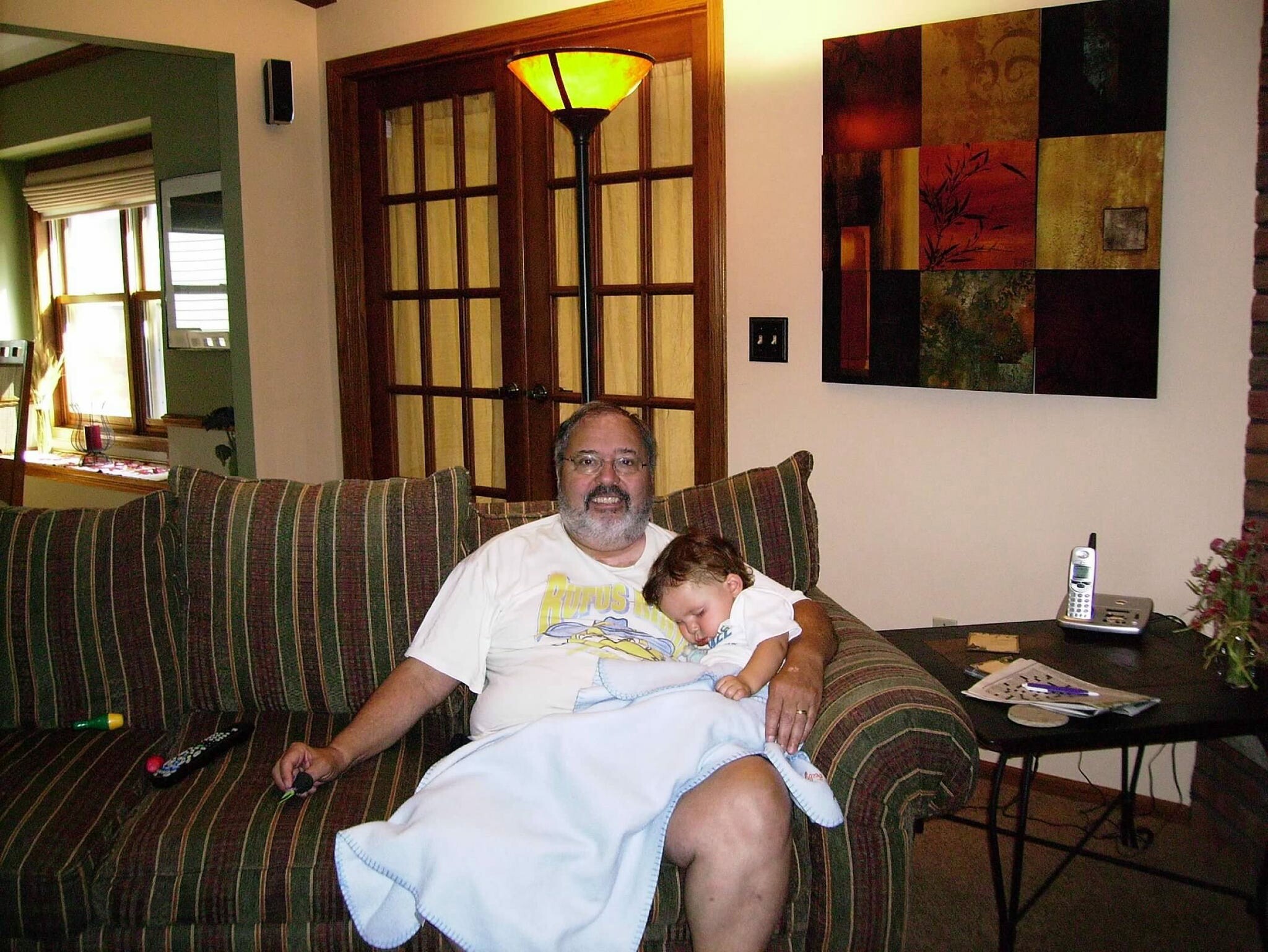 Man and child resting on a striped sofa in a living room, with a floor lamp, wall art, and a cordless phone on a side table nearby