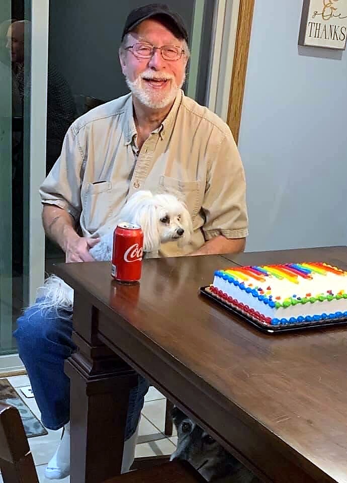 Smiling elderly man with cap sits at a table with a small white dog on his lap, a red Coca‑Cola can, and a rainbow-frosted cake nearby.