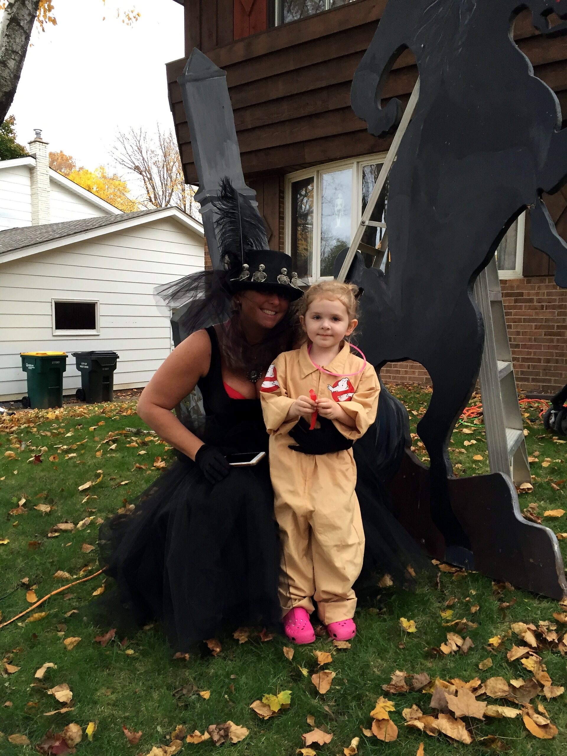 A smiling woman in a black witch costume with a tall feathered hat kneels beside a young girl in a beige ghost-themed costume outdoors on a fall day with fallen leaves and a large dark sculpture behind them.