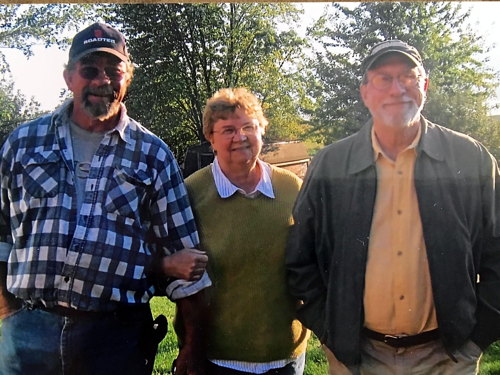 Three adults pose together outdoors, smiling at the camera with trees in the background.