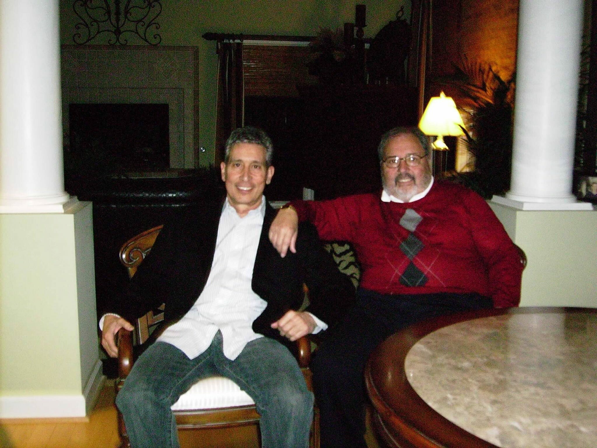 Two men sit on a porch at night, smiling for the camera; one in a dark blazer, the other in a red argyle sweater with a white shirt collar visible beneath.