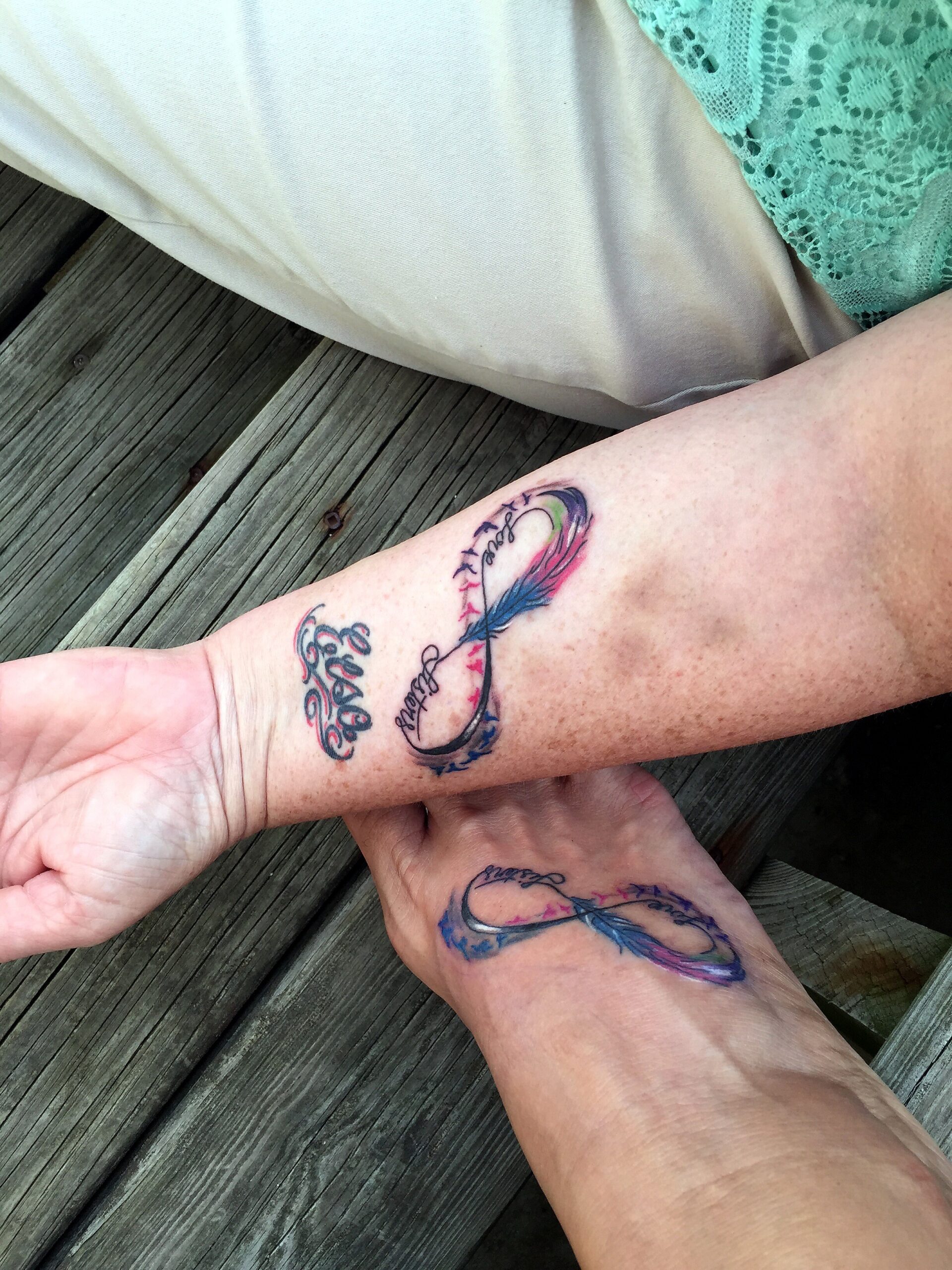 Forearm tattoo with a multicolored feather and cursive script on weathered wooden planks. A teal lace garment is visible nearby.