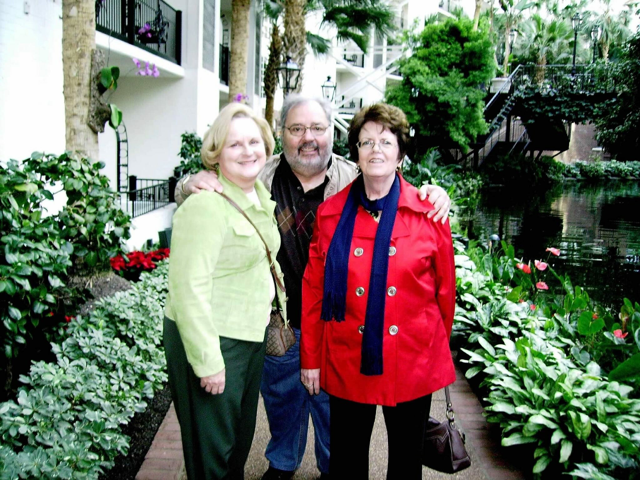 Three adults pose together outdoors in a lush courtyard with plants and a path, smiling at the camera (woman in green jacket left, man in middle, woman in red coat right).