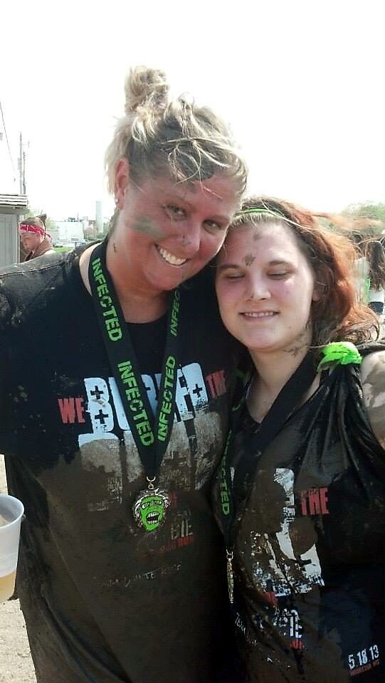 Two women at a muddy outdoor obstacle race posing together, wearing matching black shirts and green lanyards that say INFECTED, smiling at the camera.