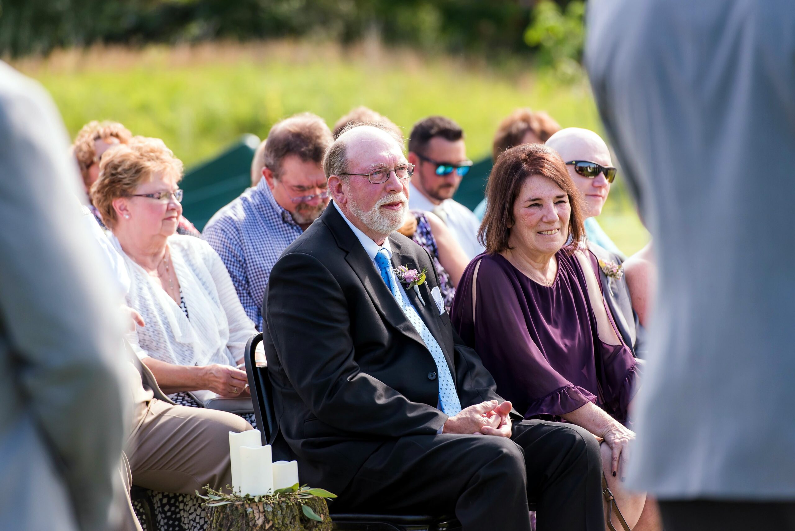 Older man in a suit and blue tie seated among wedding guests at an outdoor ceremony, with candles and greenery in the foreground.
