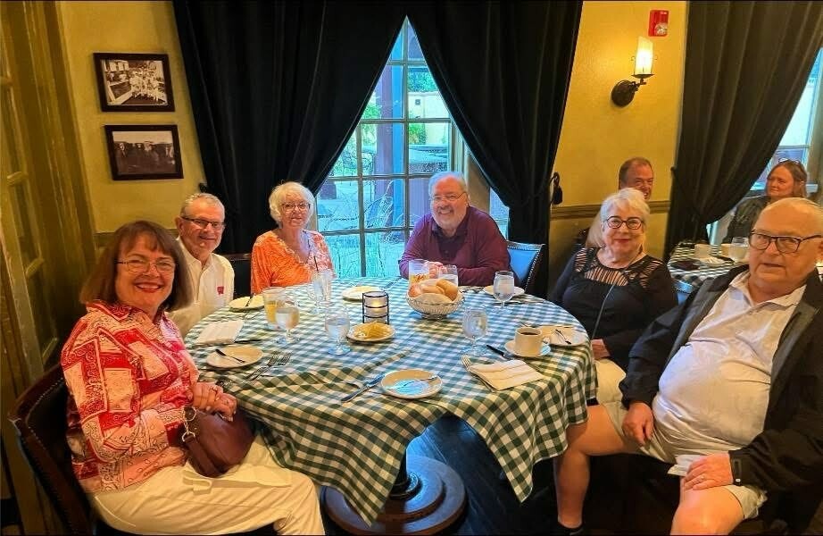 A group of older adults smiling at the camera around a round table in a cozy restaurant with a green-checkered tablecloth.