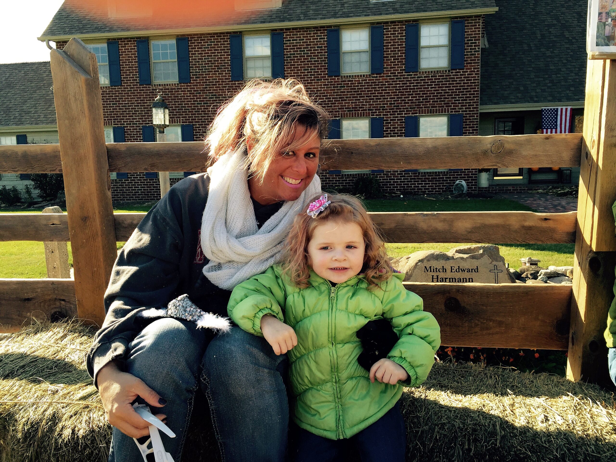 Mother and child sitting on a wooden fence outdoors, brick house with blue shutters in the background; a US flag is visible to the right.