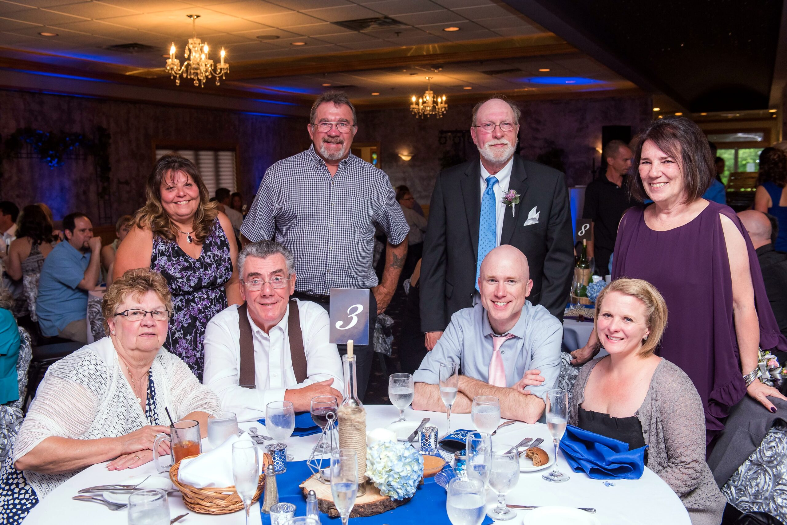 Group of smiling guests posing at a banquet table with blue decor and chandelier-lit room in the background.