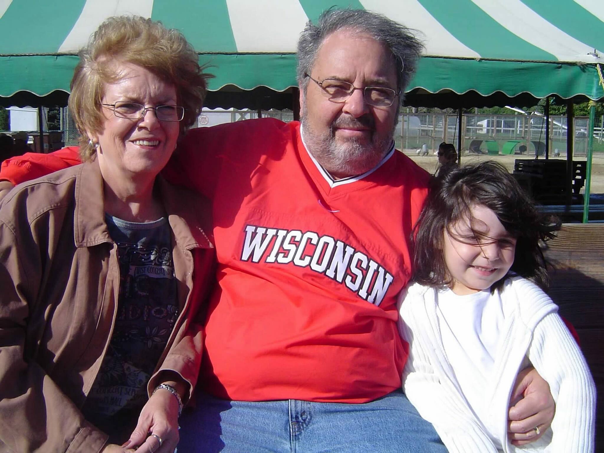 Three people pose for a selfie outdoors: an older woman left, a man in a Wisconsin shirt center, and a smiling child on the right.