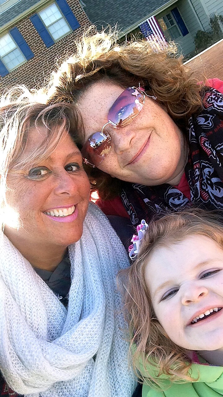 Three smiling people posing for a selfie outdoors in front of a brick house with an American flag, including two adults and a child.