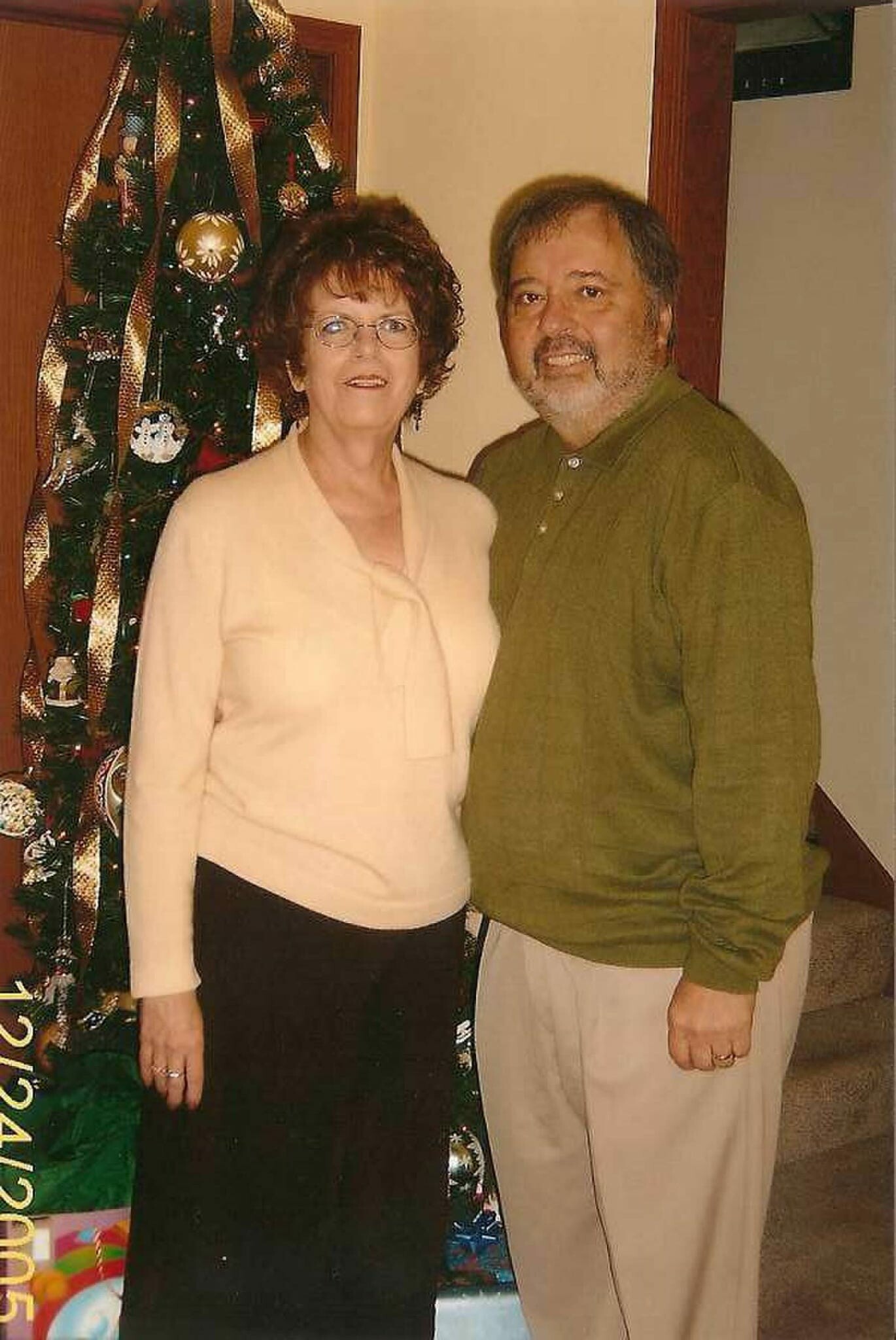 Older couple posing indoors by a tall Christmas tree with gold ribbons and ornaments.