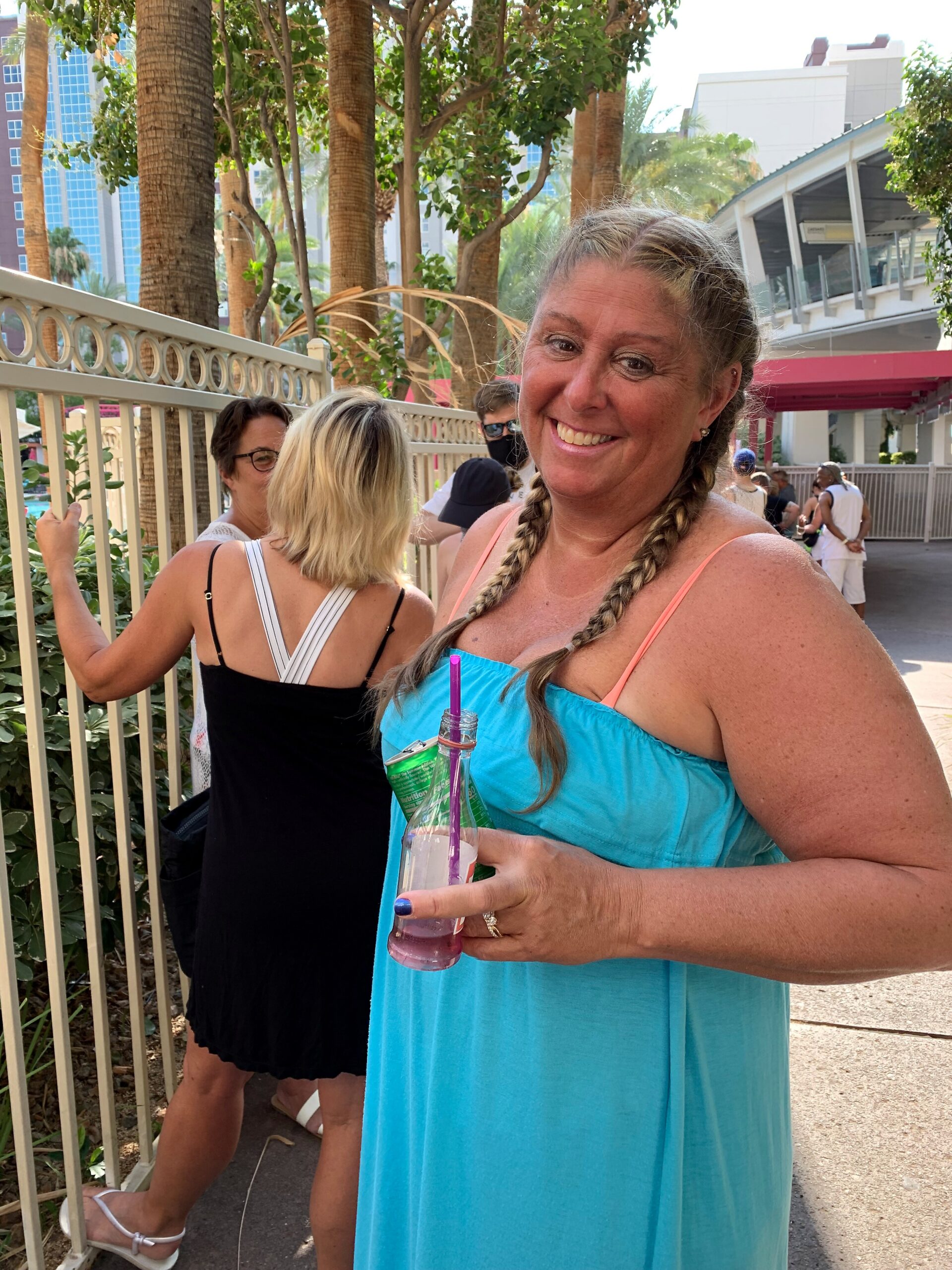 Smiling woman in a turquoise dress holding a bottle with a purple straw at an outdoor event.
